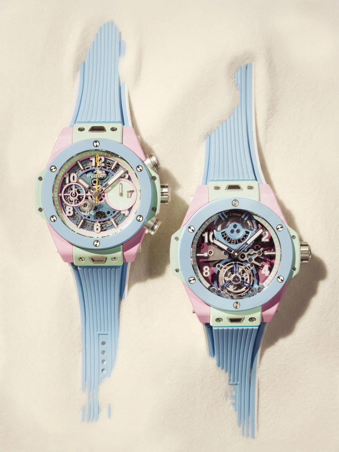 Two Hublot Big Bang Unico watches in pastel pink, mint green, and light blue with skeleton dials, on white sand.
