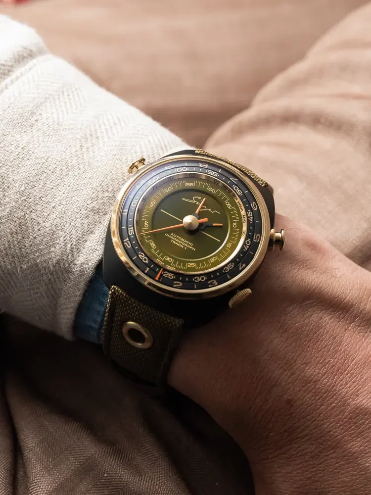 Wrist wearing a Singer Reimagined Track 1 Emirates Edition watch. Olive green dial, gold bezel, black case, and green canvas strap.