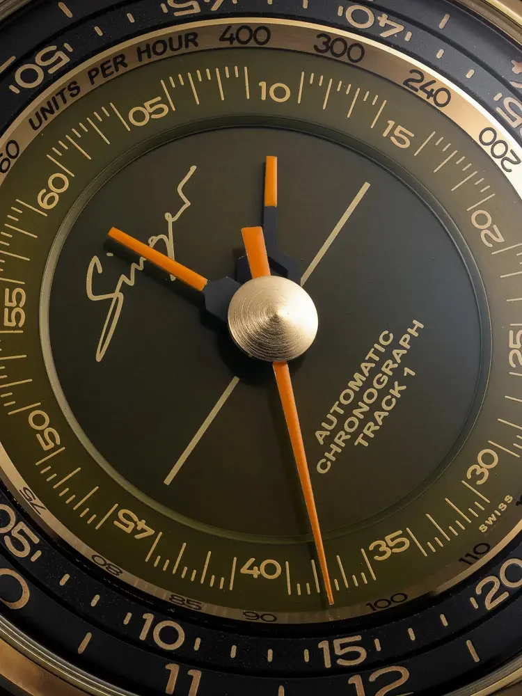 Close-up: Singer Track 1 watch dial. Olive green with gold tachymeter scale, logo, and orange chronograph hands.
