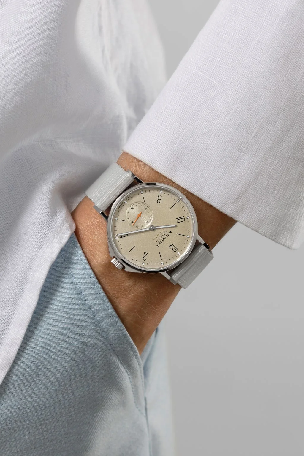 Wrist wearing a Nomos Ahoi Neomatik with light beige sand dial, grey strap, orange small seconds, white shirt.