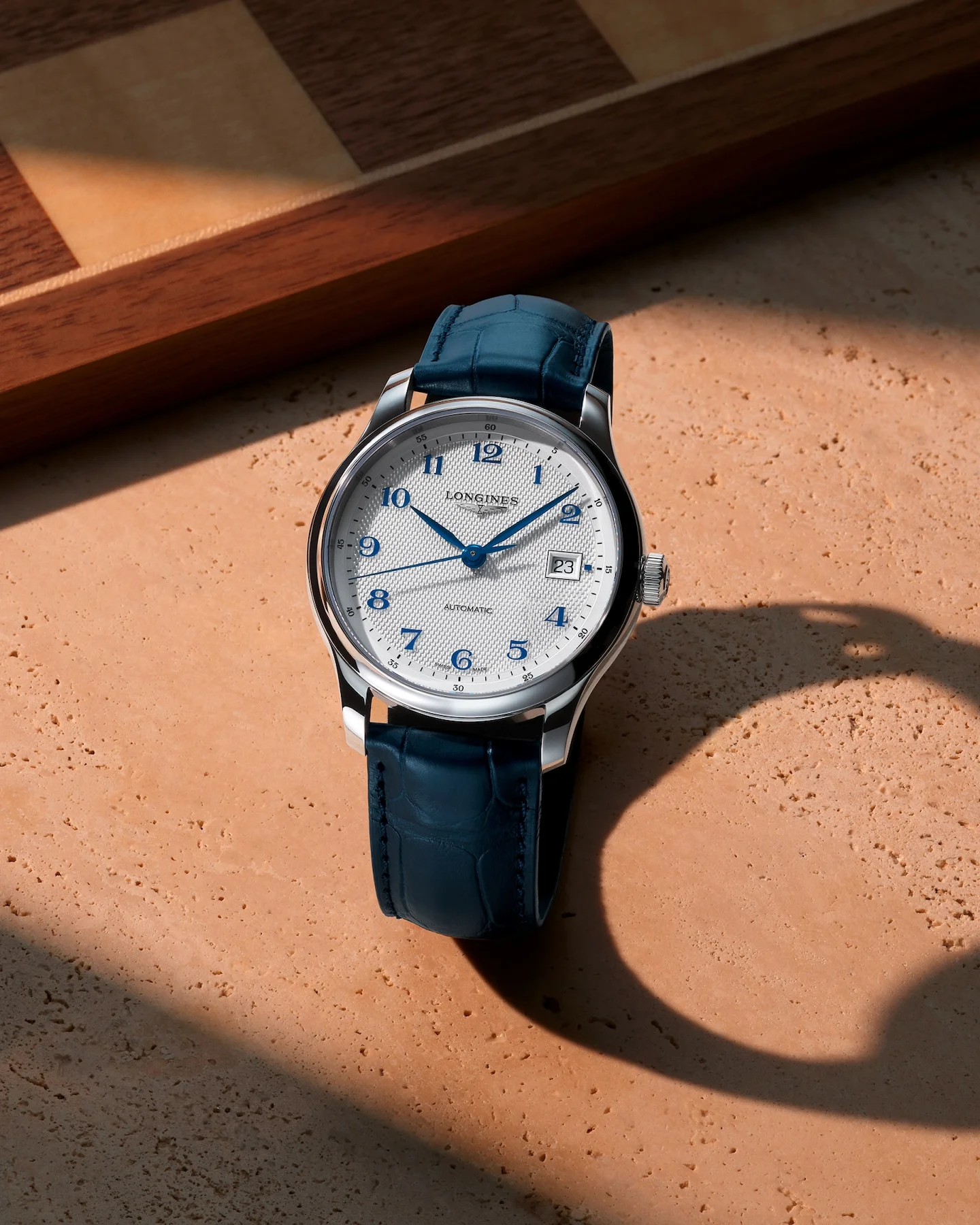 Longines watch: silver case, white patterned dial, blue Arabic numerals & hands, date (23), blue alligator strap, on stone & wood.