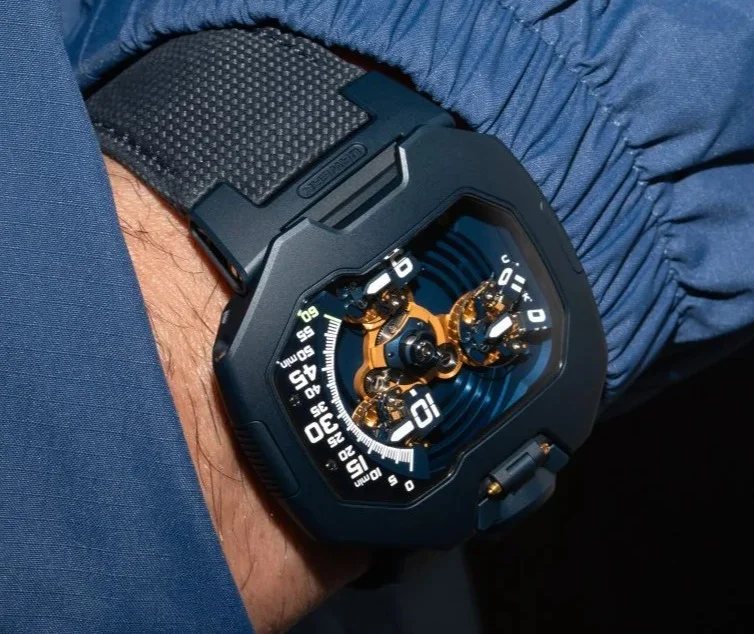 URWERK UR-120 watch on a wrist with blue jacket. Features a black case, intricate gold mechanism, and black textured strap.