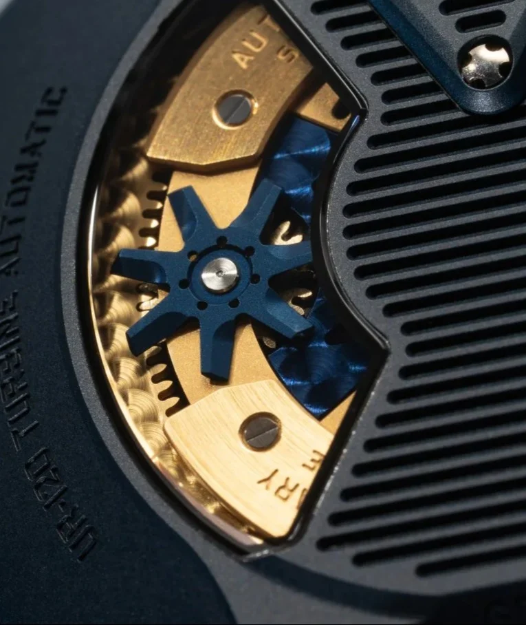 Close-up of URWERK UR-120 watch mechanism: golden gears, blue turbine, and "TURBINE AUTOMATIC" engraving on dark case.