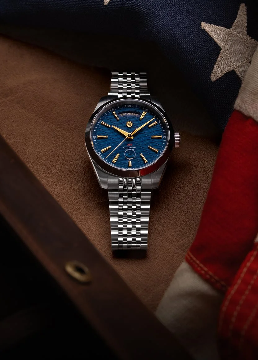 A wristwatch with a blue, wavy dial and steel bracelet resting on a leather surface near an American flag.