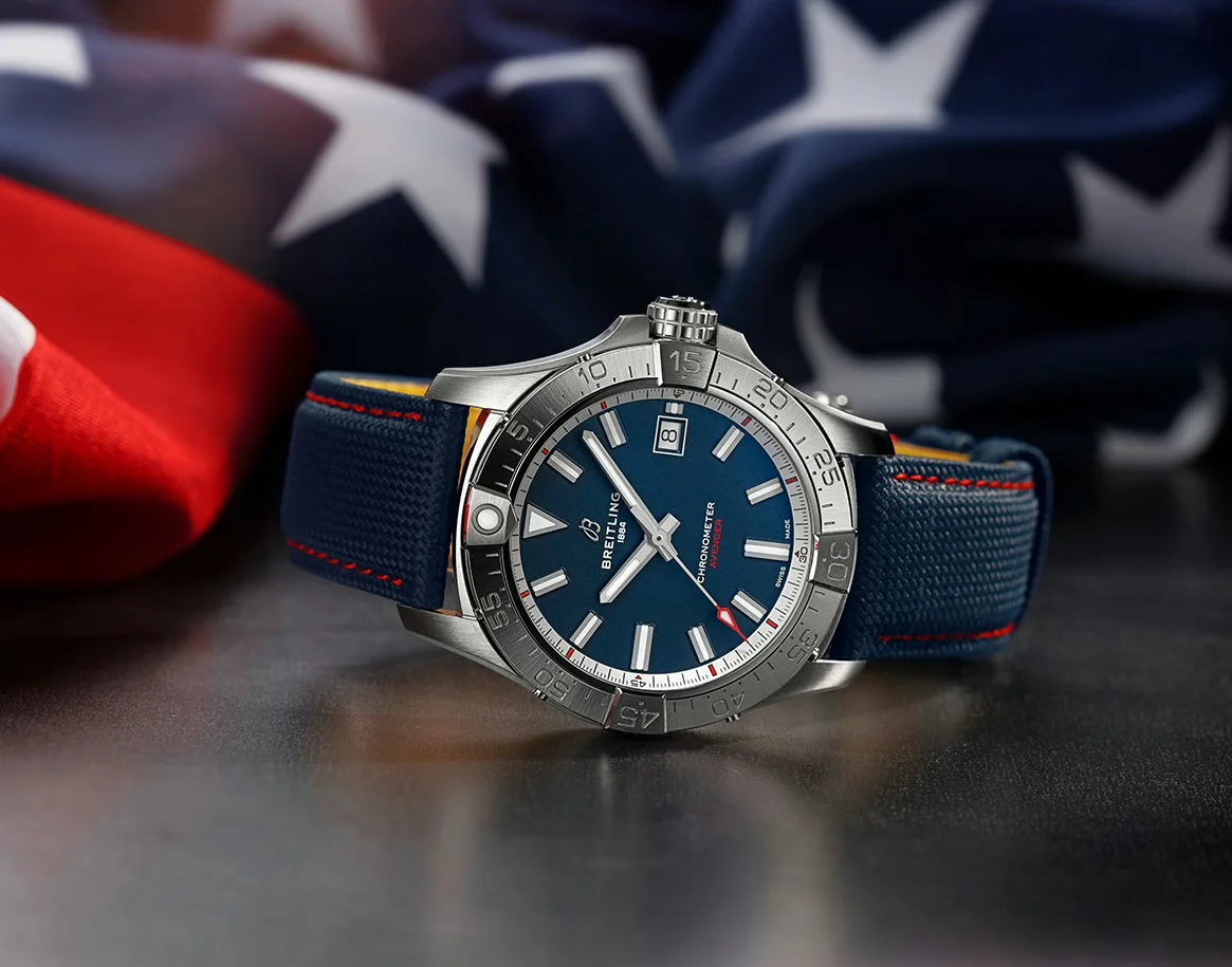 Breitling Avenger with blue dial, canvas strap with red stitching, on dark background with US flag.