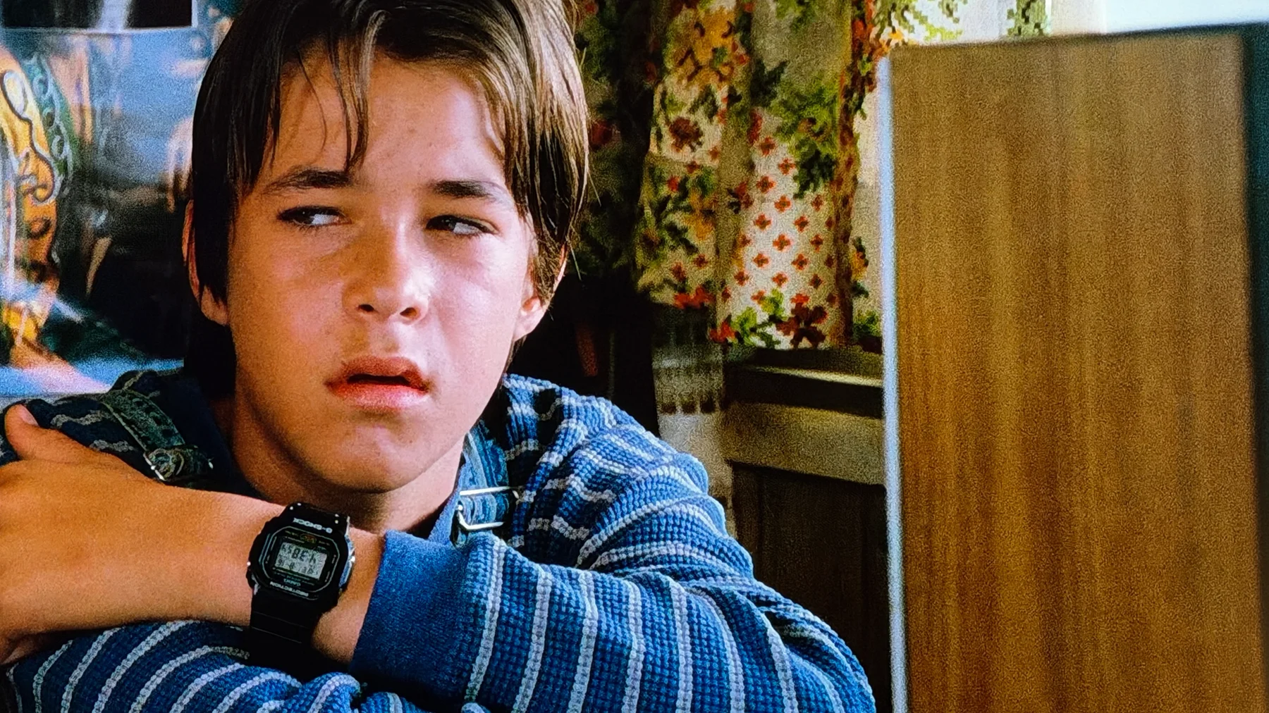 Young boy in blue shirt with a black G-Shock watch on his left wrist, looking right. Floral curtain & wooden furniture.