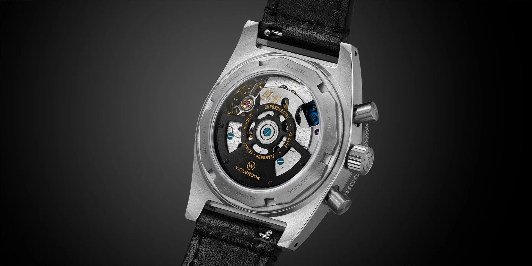 Wolbrook steel watch, exhibition caseback reveals intricate chronograph movement with gold accents, blue screws, and purple jewels. Black leather strap.