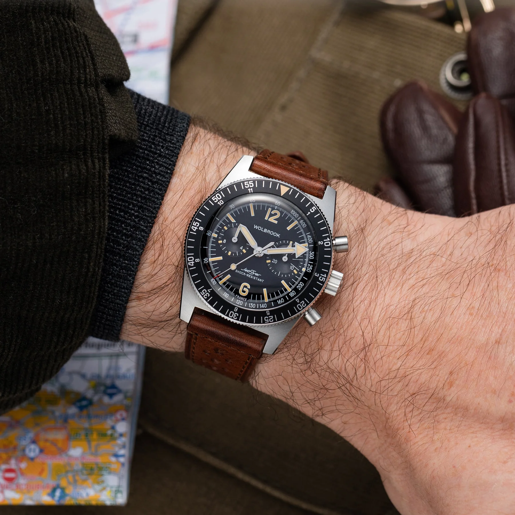Wrist shot of a Wolbrook Jet-Timer watch with steel case, black dial, brown leather strap, over a map & glove.