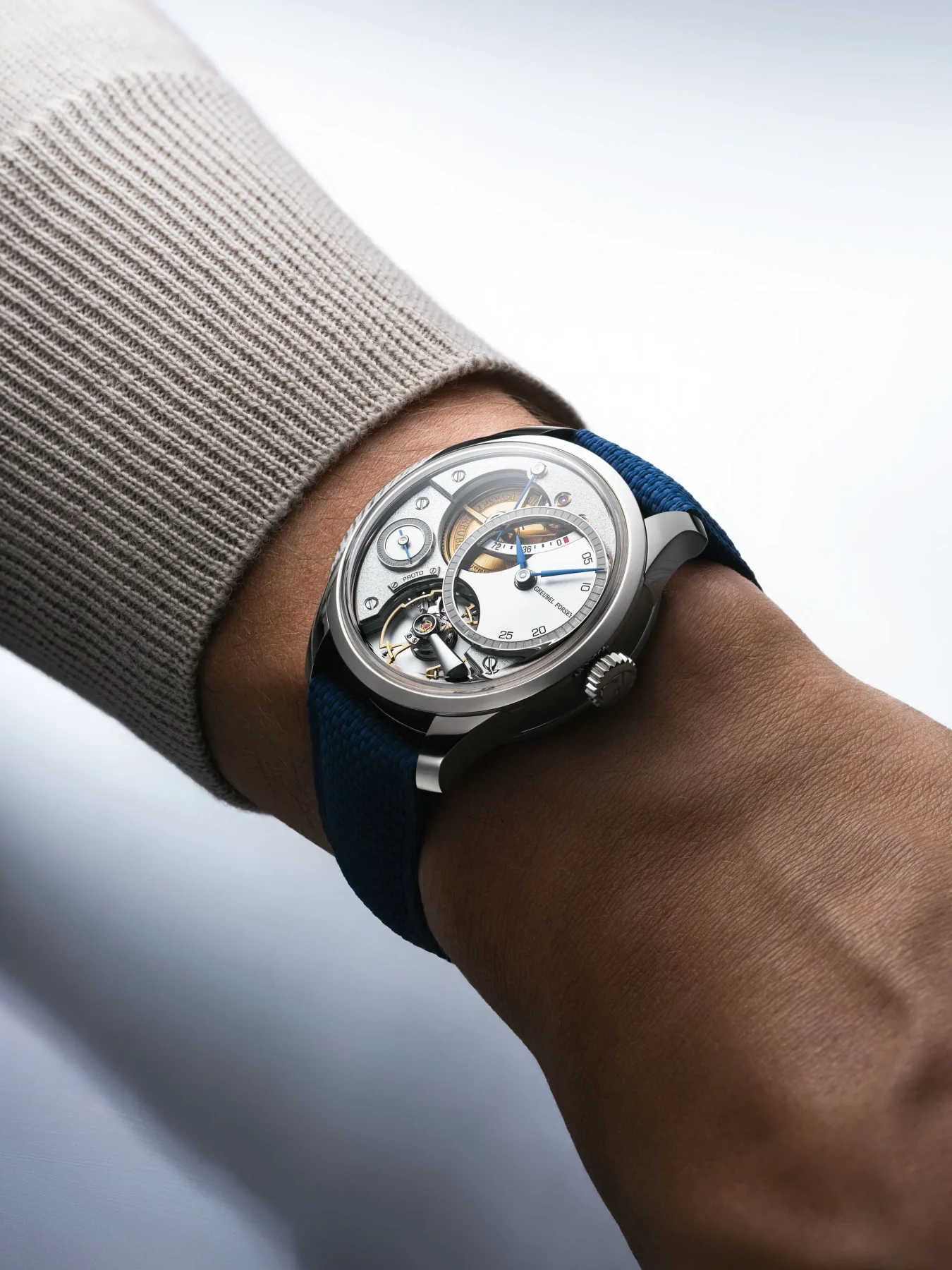 Wrist with a Greubel Forsey watch: openworked dial, silver case, blue strap, beige knitted sweater sleeve.