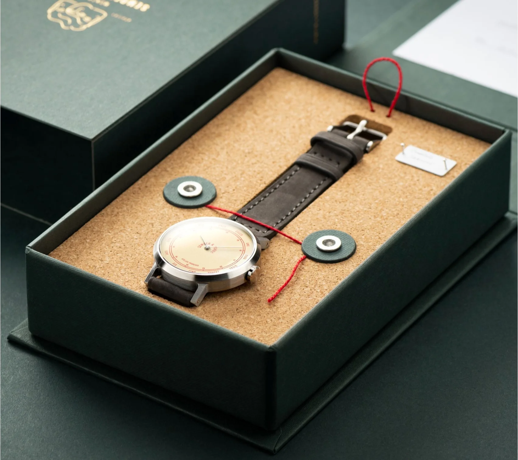 Luxury watch in open green box. Silver case, light dial, brown leather strap secured by red string in cork bed.