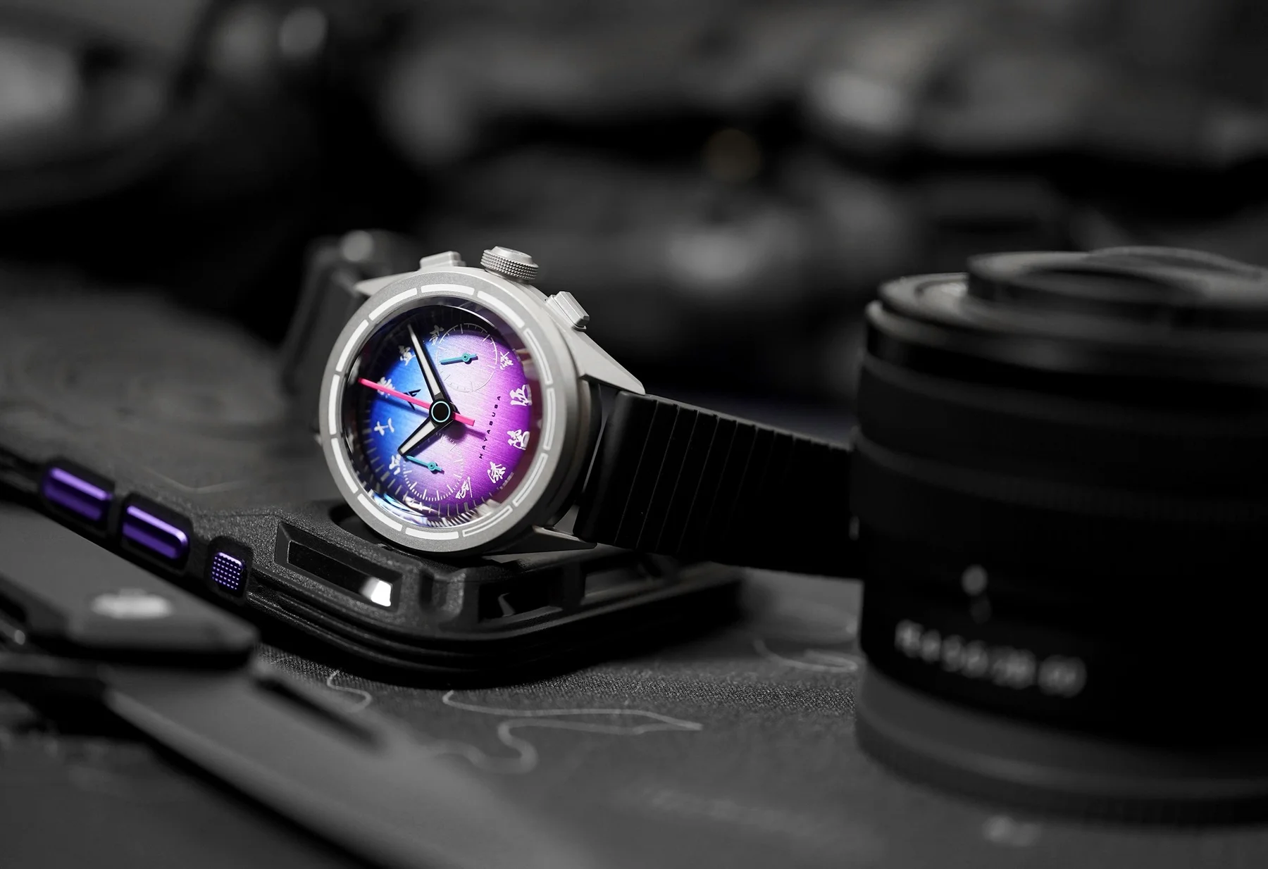 Vibrant blue-purple Hayabusa chronograph watch, silver case, black rubber strap, on a dark surface with phone case & lens.