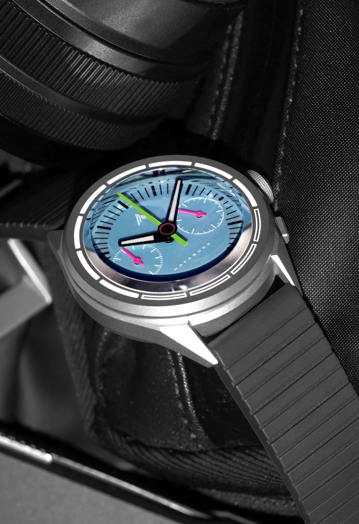 Angular silver HAYABUSA watch with light blue dial, green/black hands, pink sub-dials, black ridged strap on dark fabric.