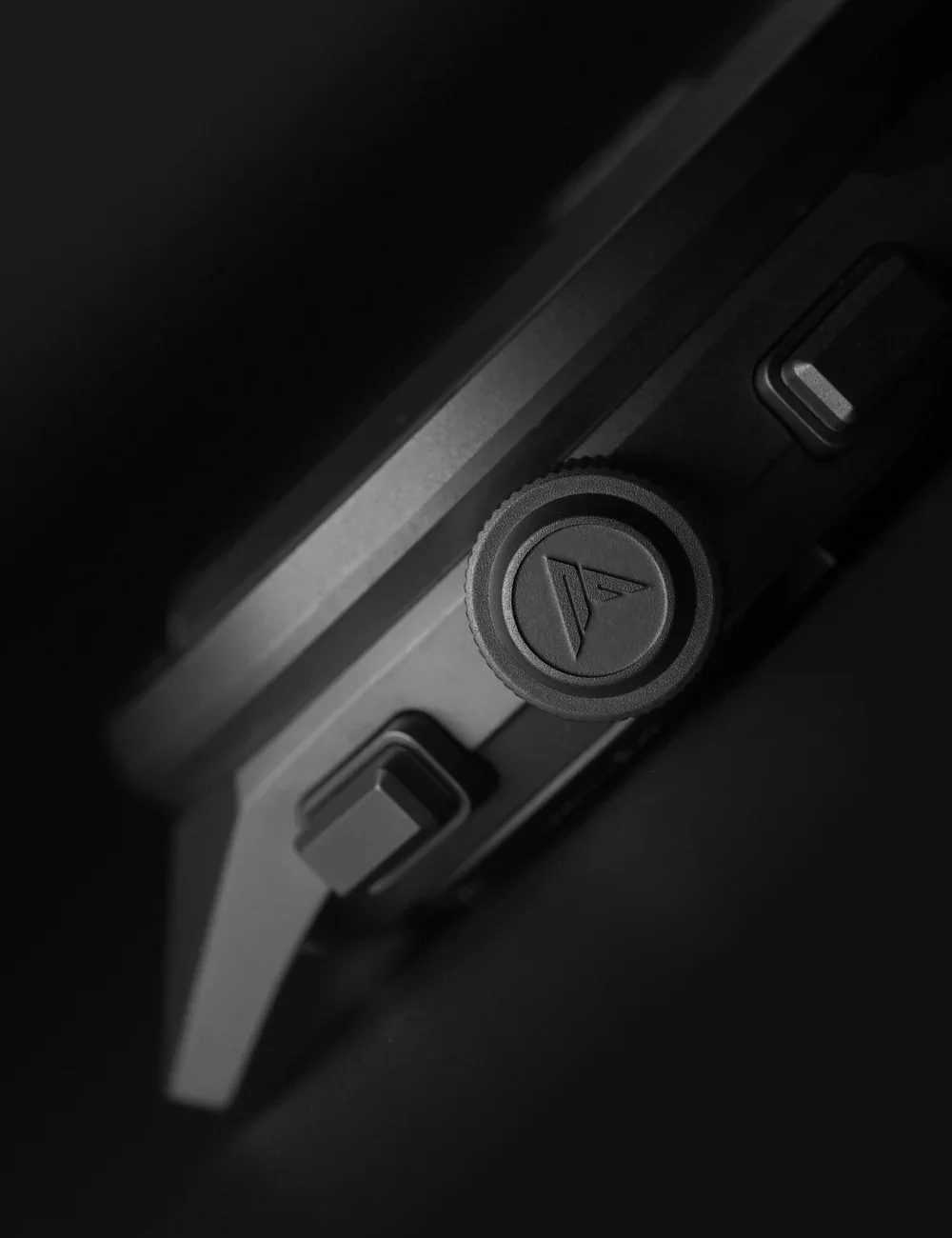 Angled close-up of a black luxury watch case, showing a textured crown with a stylized logo and two rectangular pushers.