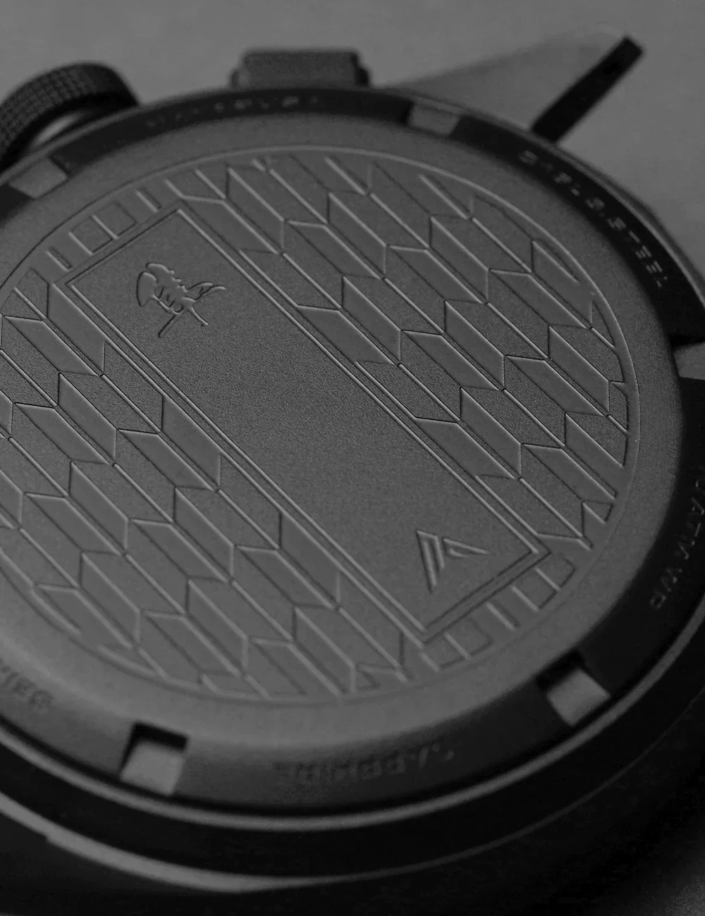 Close-up of a matte dark watch case back with engraved dragon figure, 'A' logo, and geometric Art Deco pattern.