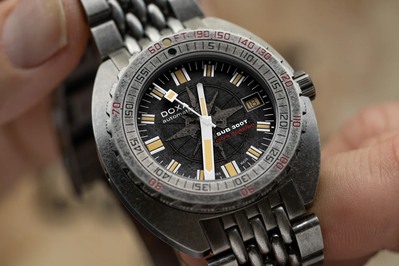 Close-up of DOXA SUB 300T Clive Cussler watch with weathered gray bezel & black compass dial, held by fingers.