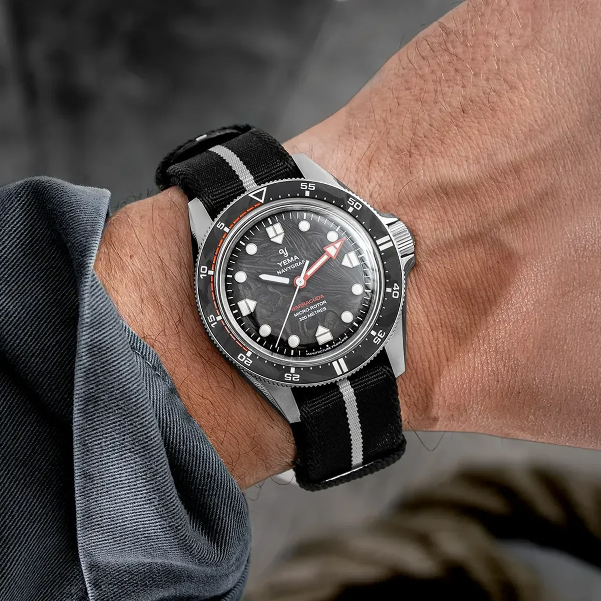 Man's wrist wearing a Yema Navygraf Barracuda diver watch. Steel case, black textured dial with orange details, black/grey NATO.