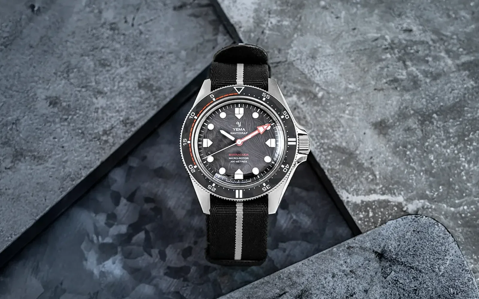 Yema Navygraf Barracuda watch on dark textured background. Silver case, black textured dial, red minute hand, black/white NATO strap.