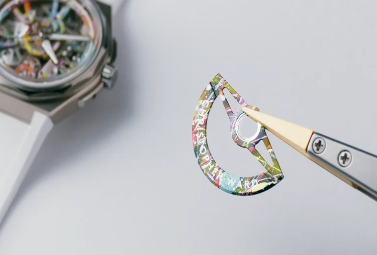 Tweezers hold a paint-splattered Christopher Ward rotor. Blurred skeleton watch with matching colorful dial in background.