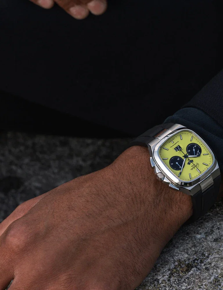 Dark arm wearing a cushion-shaped silver watch with a vibrant yellow dial, black subdials, and black strap, on concrete.