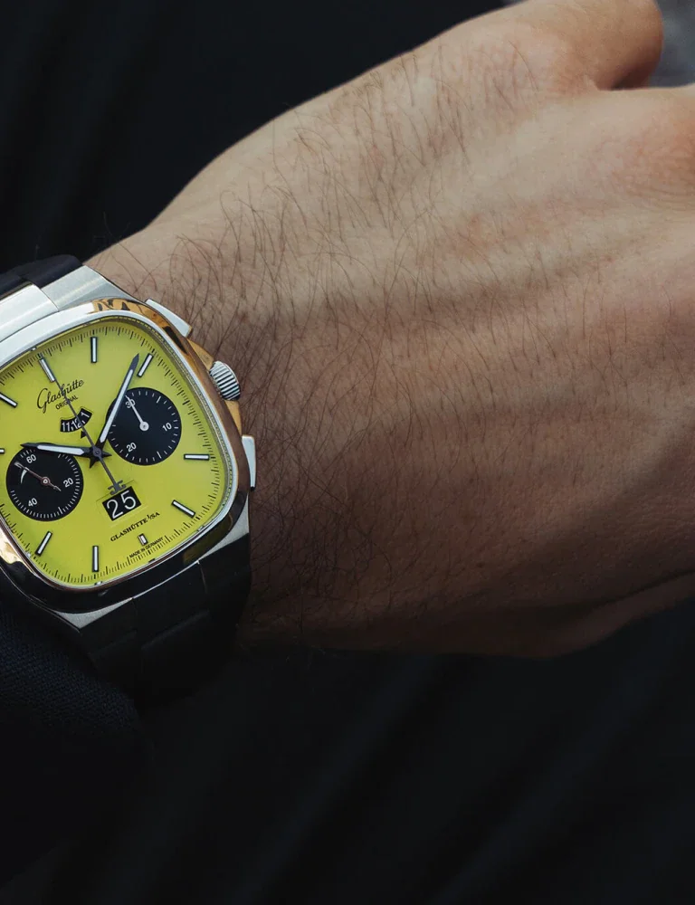 Close-up of a hairy wrist wearing a Glashütte Original chronograph. Yellow square dial, two-tone case, black strap, date 25.