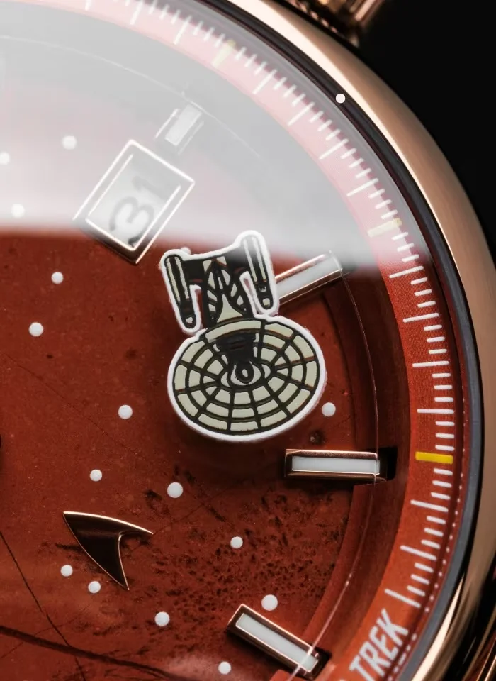 Close-up of a rose gold Star Trek watch dial. Red textured surface, Enterprise graphic, Starfleet insignia, date 31, white dots.