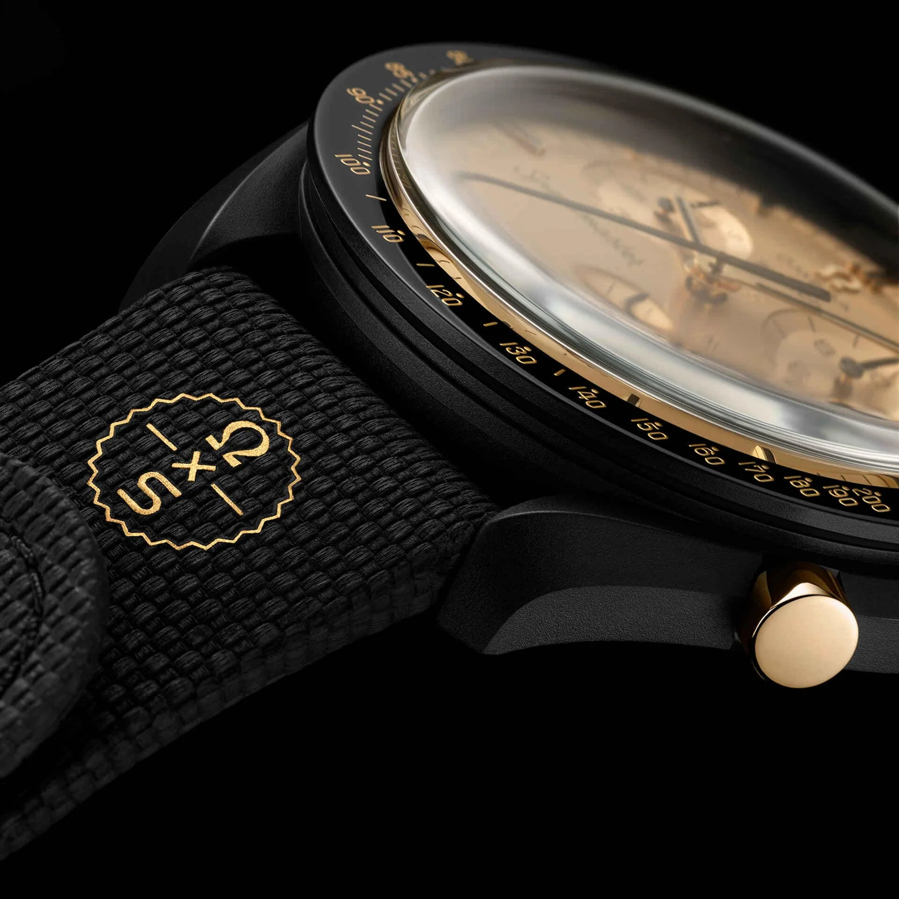 Close-up of a black watch. Black textured strap with gold 5X5 logo. Gold crown, gold tachymeter bezel, and gold-toned dial.