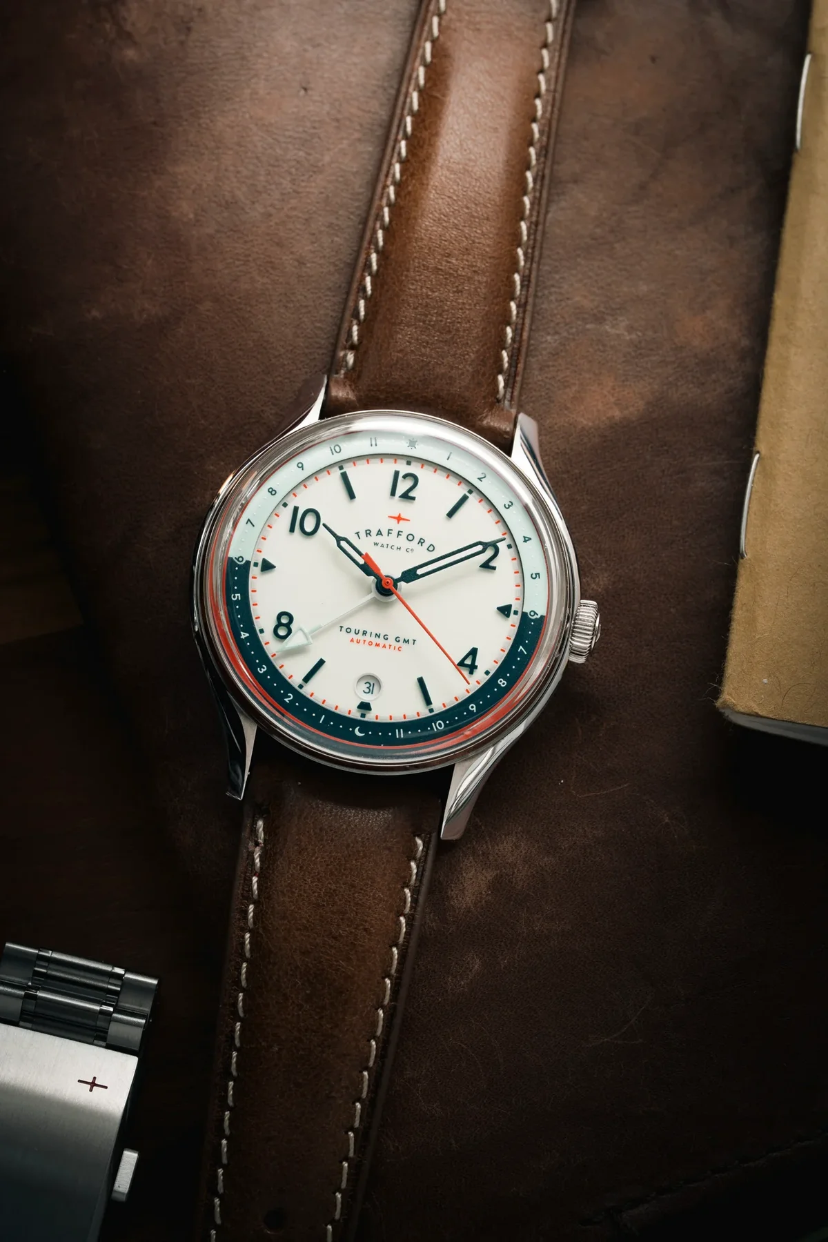 Trafford Touring GMT Automatic watch with white dial, two-tone red/blue 24-hr scale, brown leather strap, on a leather surface.