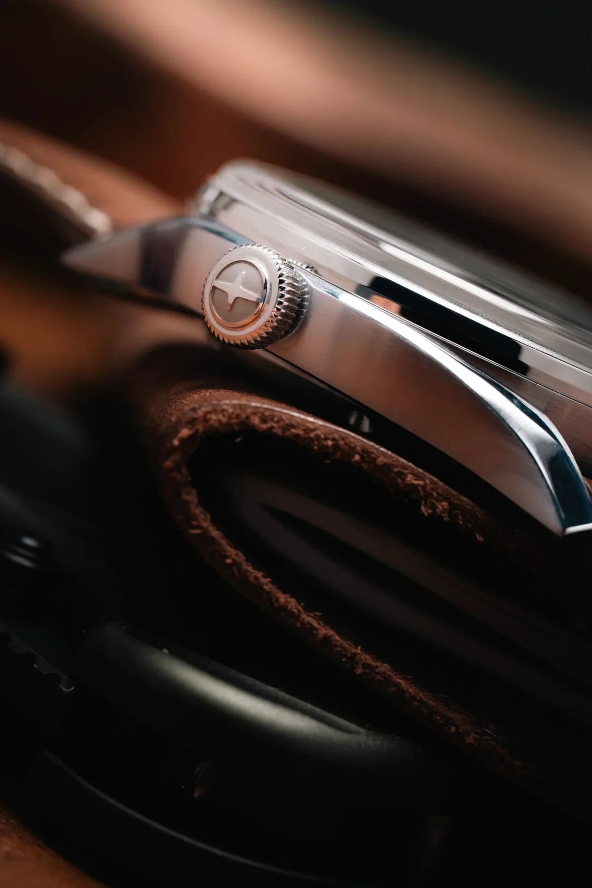 Close-up of a polished steel watch case and knurled crown with white logo, resting on a dark brown leather surface.