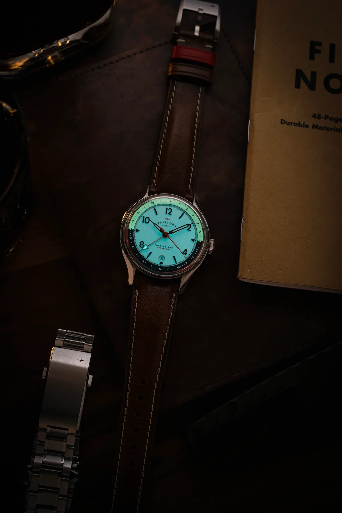 Trafford watch with glowing aqua dial and brown leather strap, on a dark desk with a metal bracelet and notebook.
