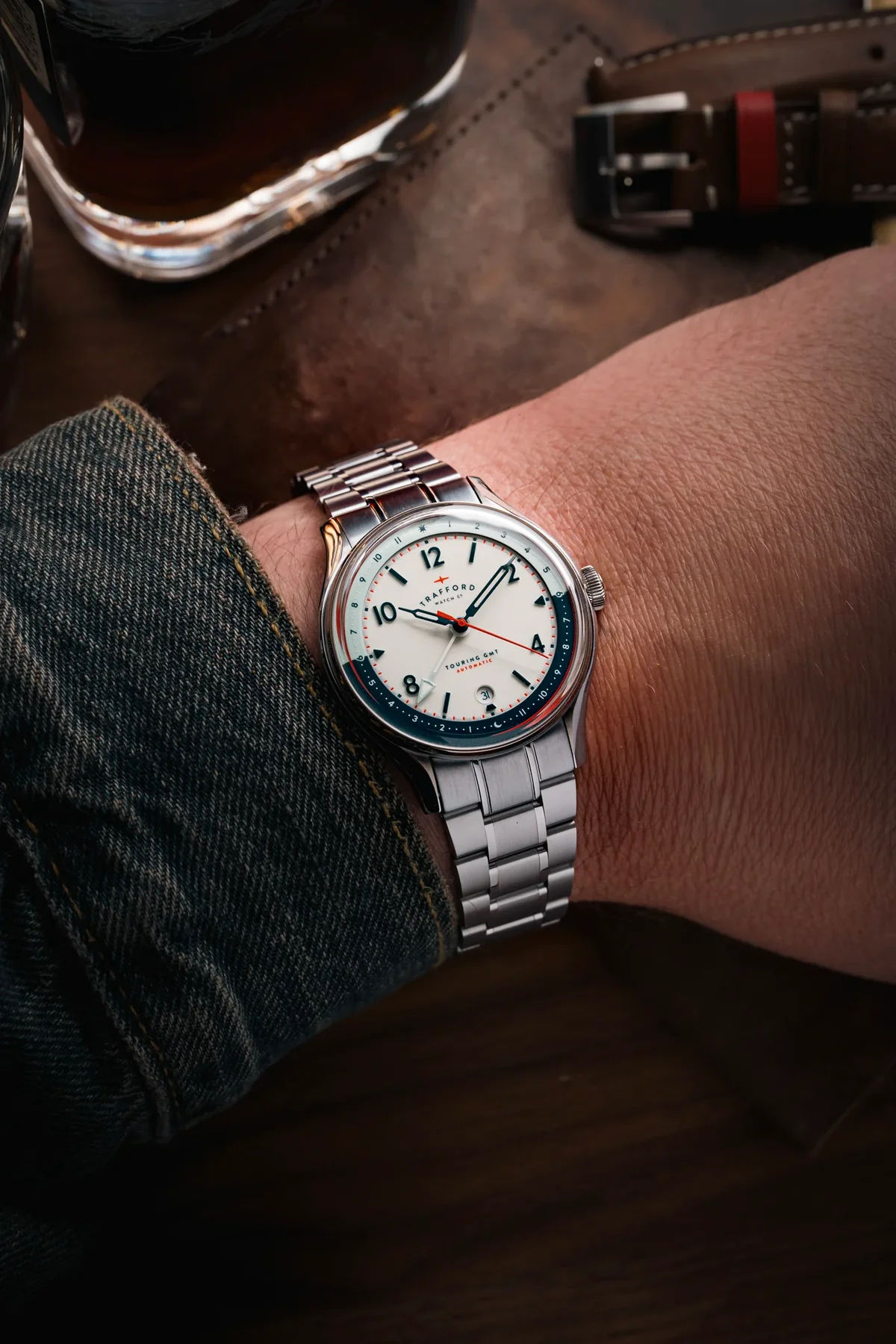 Wrist wearing Trafford Touring GMT Automatic watch, white dial with blue/red GMT ring, red GMT hand, steel bracelet. Denim, drink, leather.