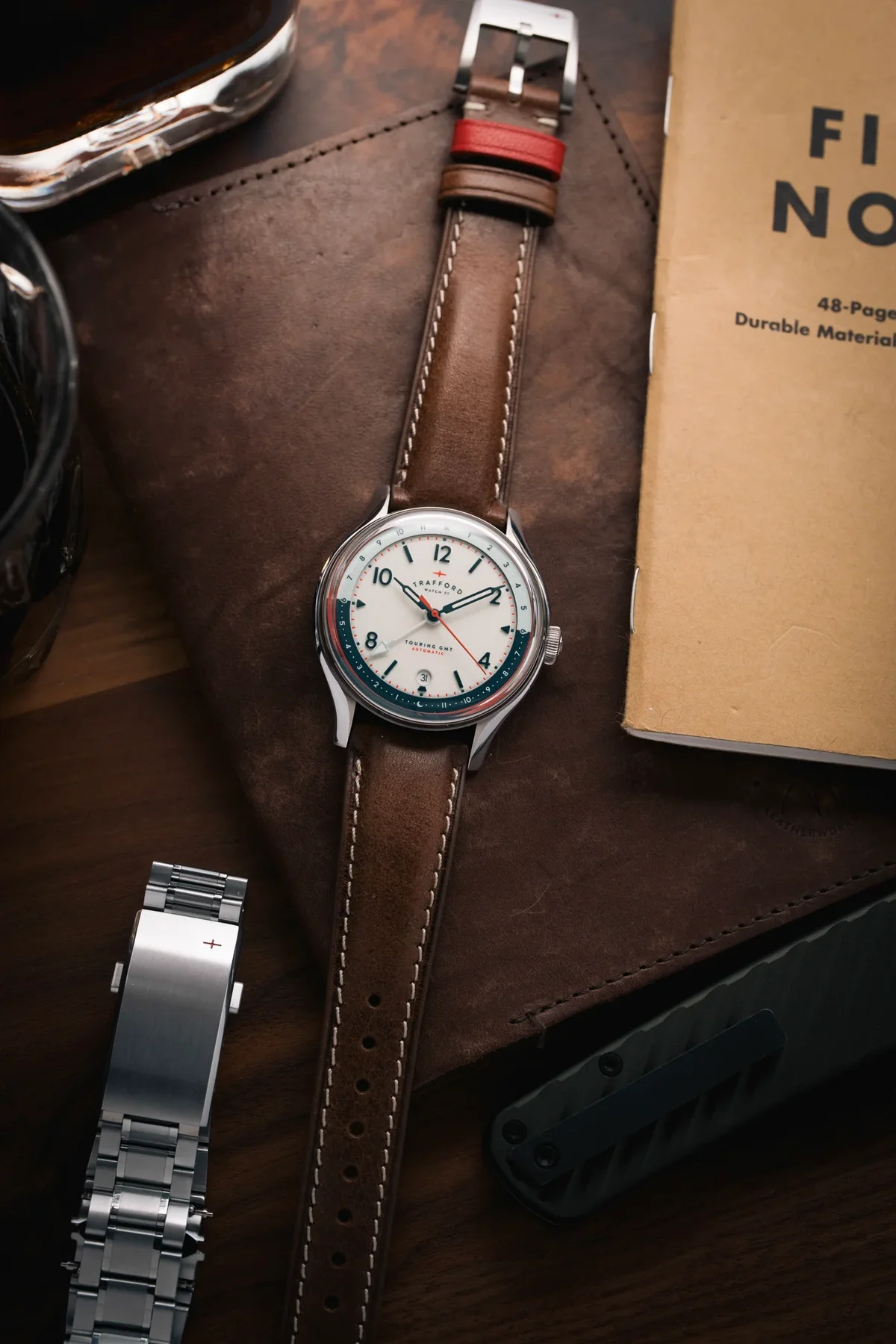 Trafford Touring GMT watch: white dial, brown leather strap with red keeper. On leather pad with notebook & other EDC items.