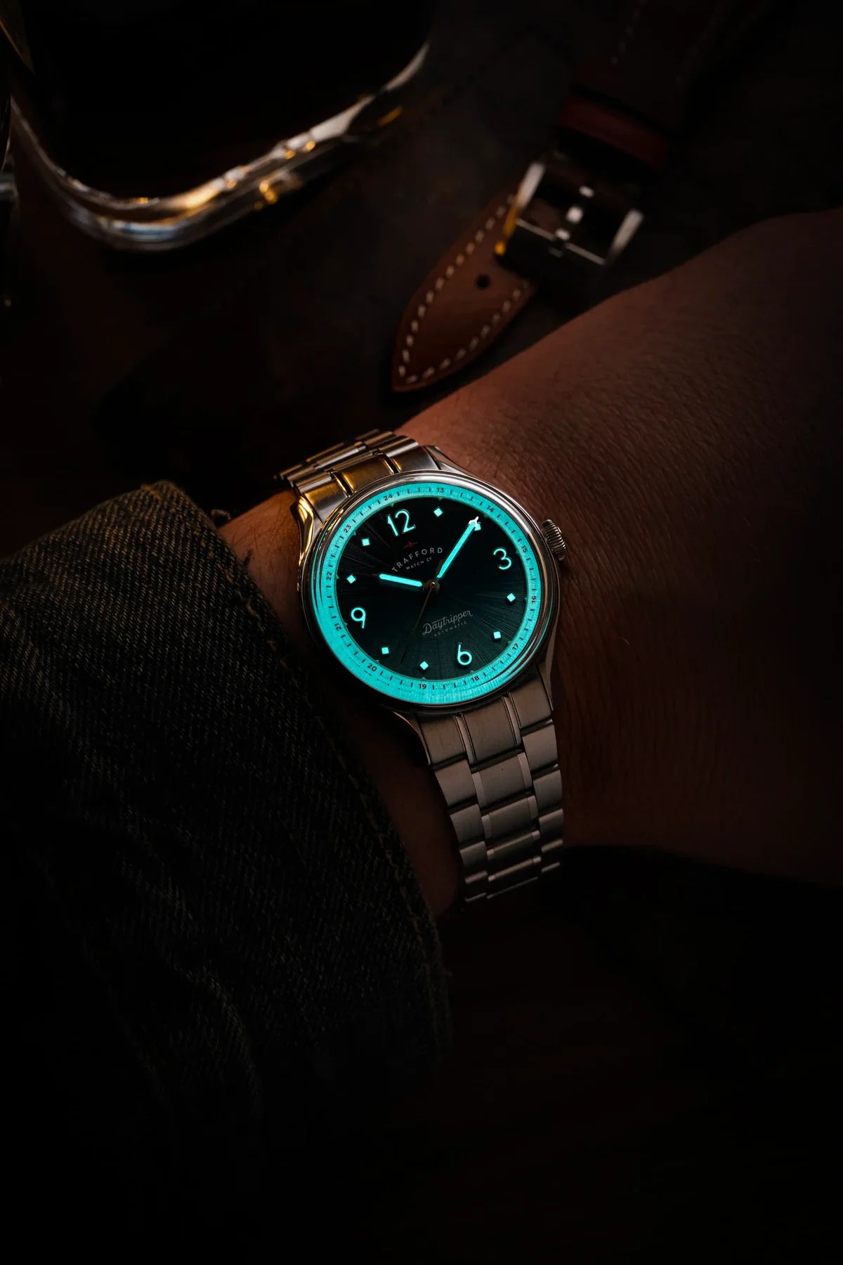 Close-up of a wrist wearing a Trafford Daytripper automatic watch. Its dark dial, markers, hands, and inner bezel glow blue.