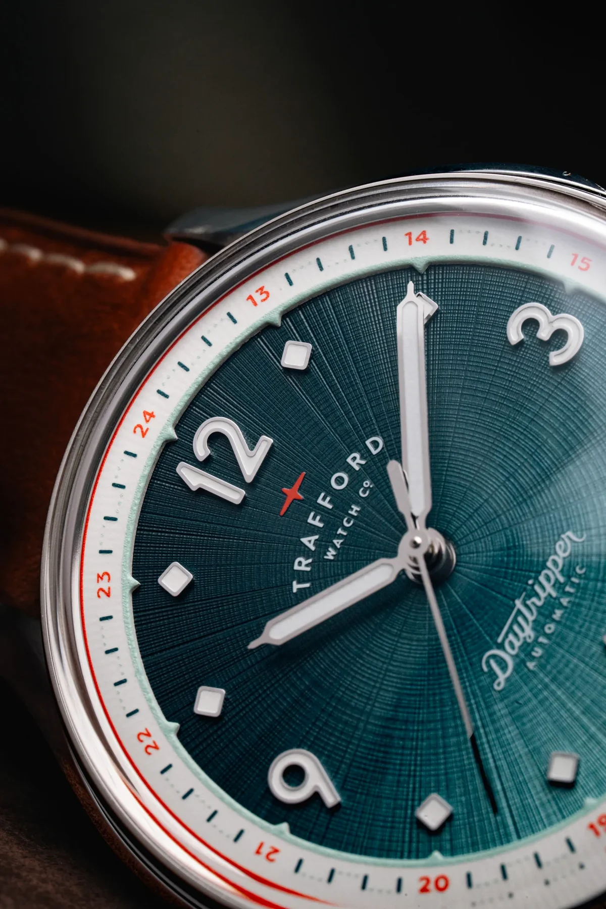 Close-up: Trafford Daytripper automatic watch. Teal textured dial, white 12, 3, 9, white hands, 24-hr scale, silver case, brown strap.