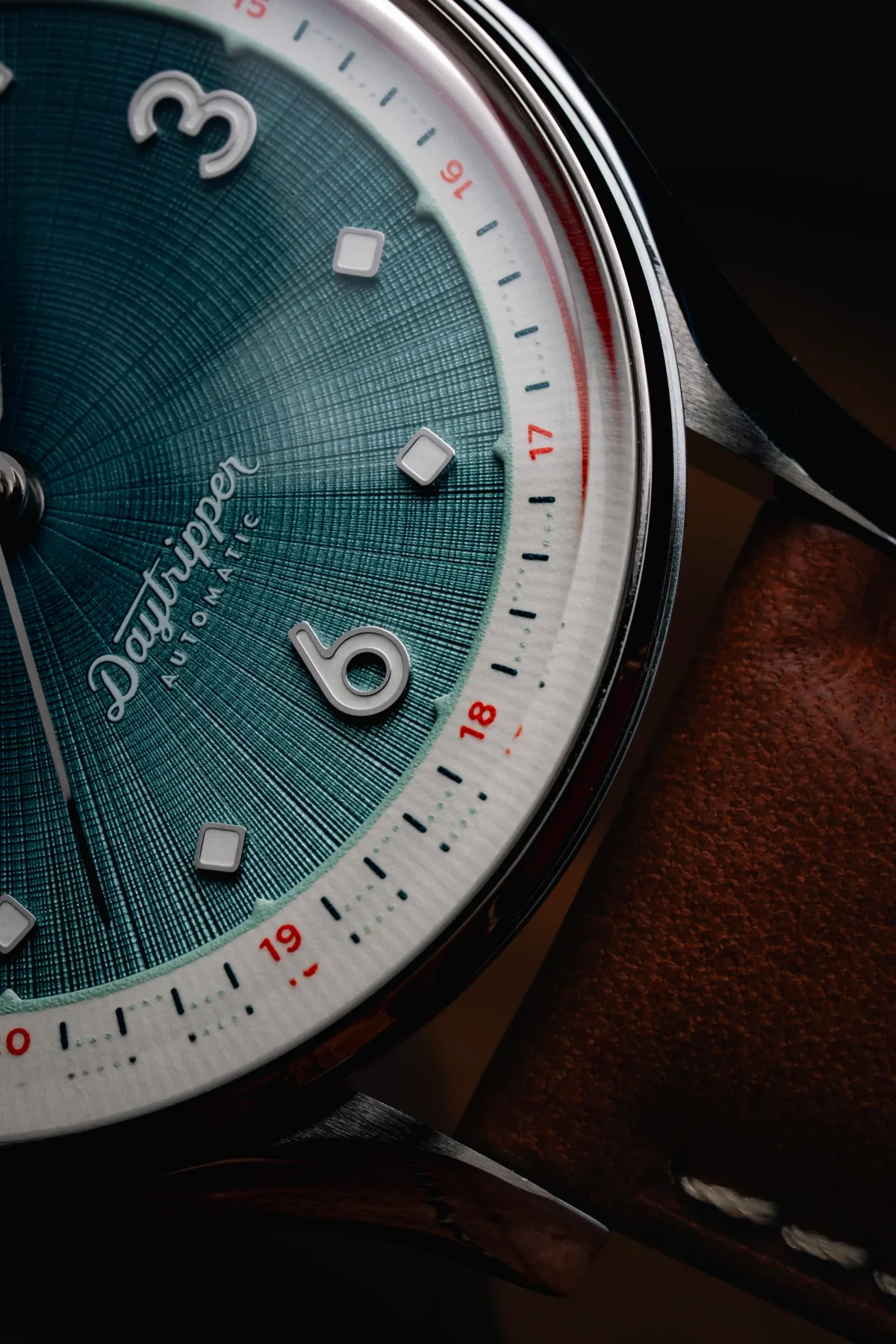 Close-up: Daytripper automatic watch. Teal textured dial, white markers, 24-hr track with red numerals, chrome case, brown leather strap.