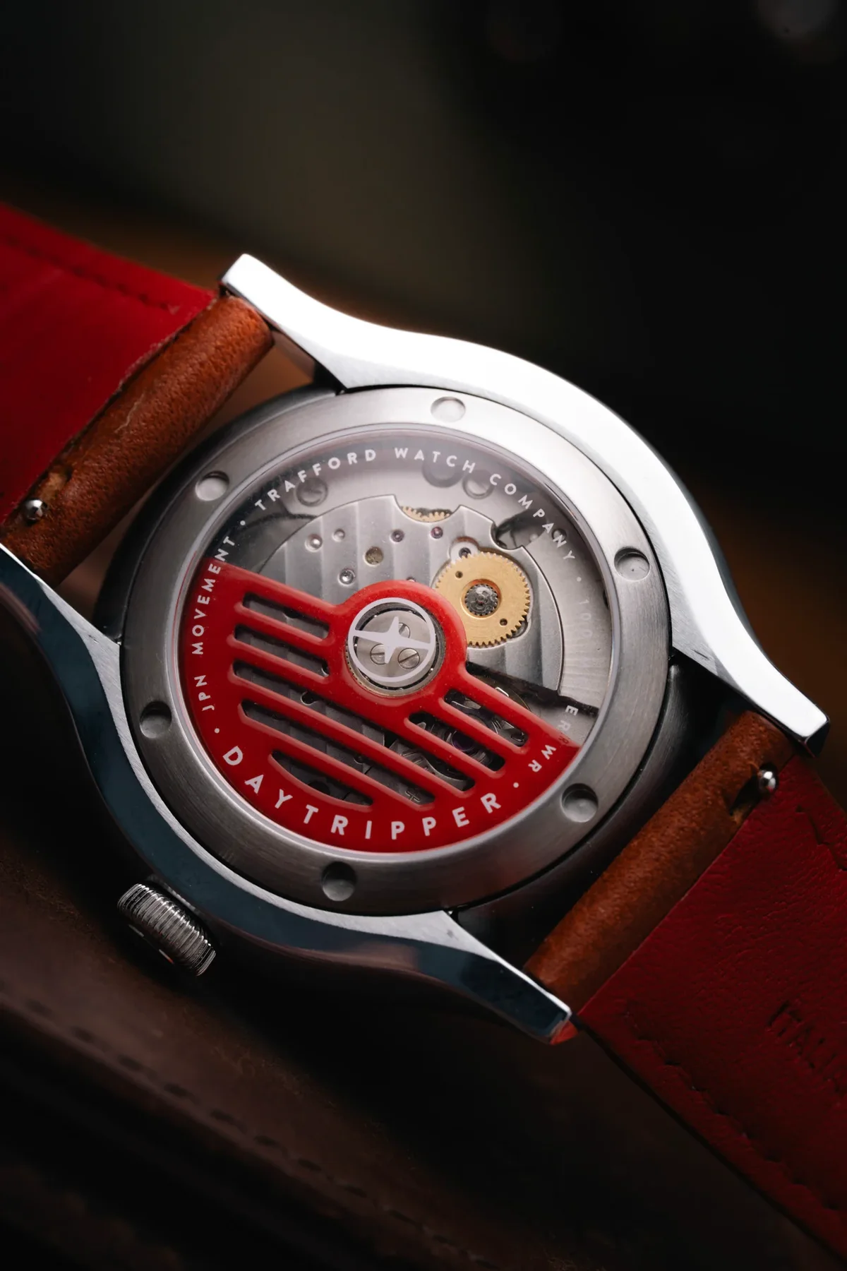 Close-up of a silver watch's exhibition case back with a red rotor, visible movement, and red leather strap. Reads 