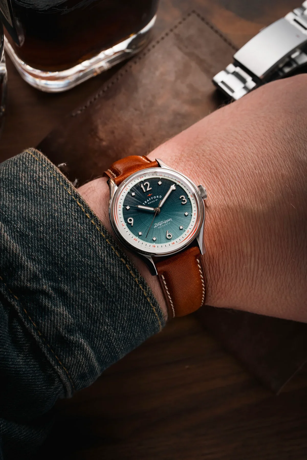 Wrist wearing a Trafford Daytripper watch with a teal sunburst dial, brown leather strap & denim cuff.
