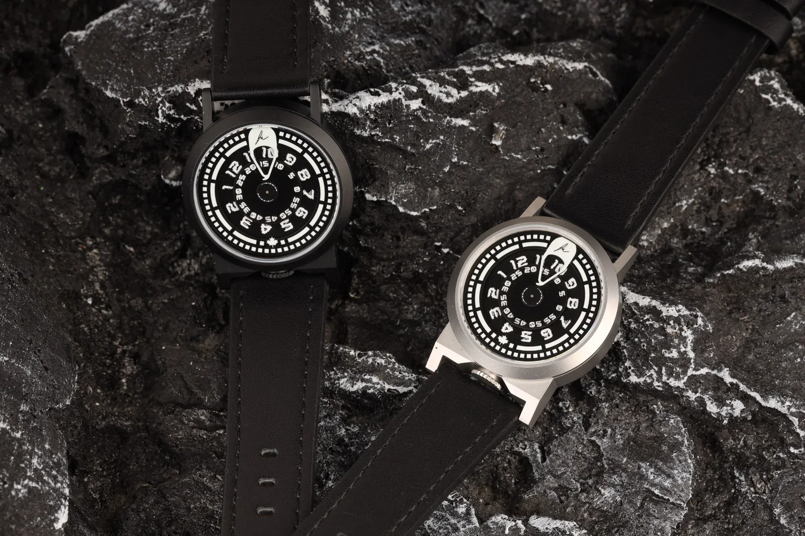 Two luxury watches, one black and one steel, with black dials and leather straps, lie on dark volcanic rock.
