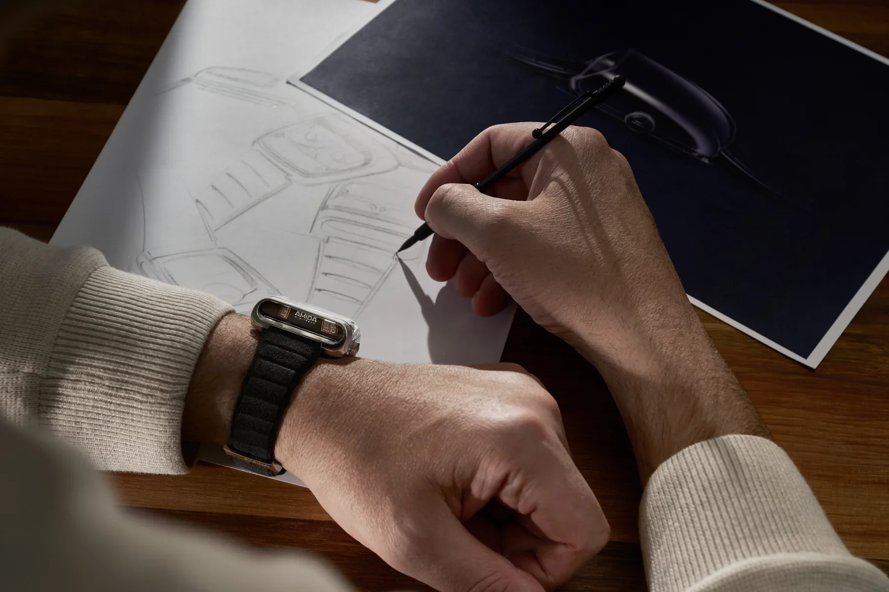 A person's hands drawing watch designs. An AMIDA DIGI-COMP watch with a silver case & black strap is on their left wrist.