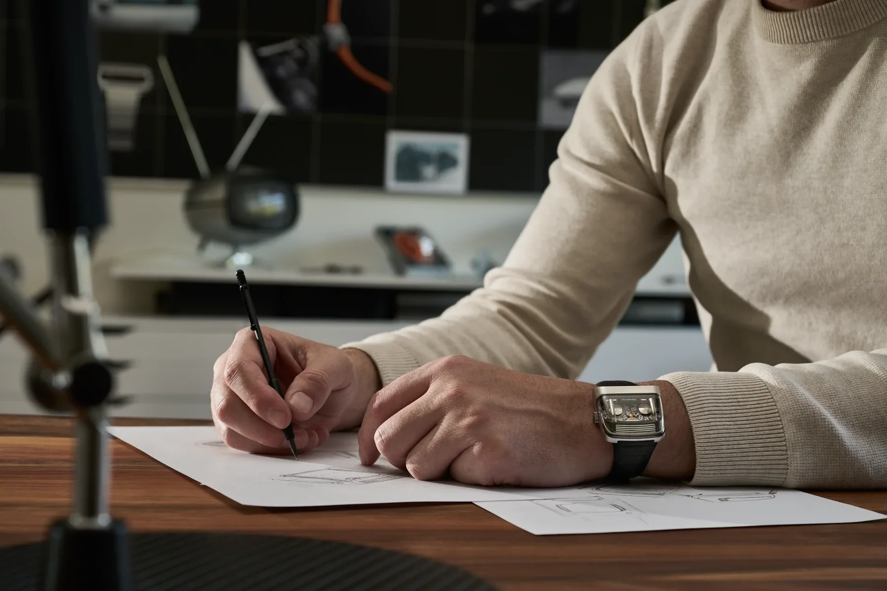 Man in beige sweater sketching watch designs at a wooden desk, wearing a distinctive ANIMA luxury watch with exposed movement.