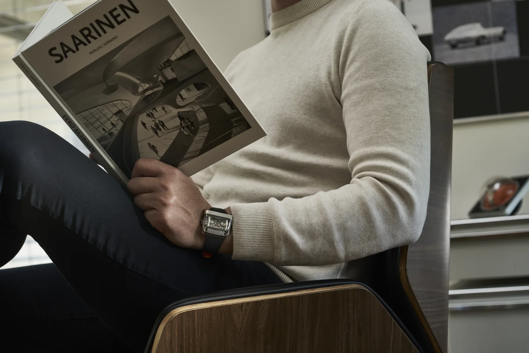 A man in a sweater reads a book, his wrist displaying a distinctive rectangular luxury watch with an intricate mechanical dial.