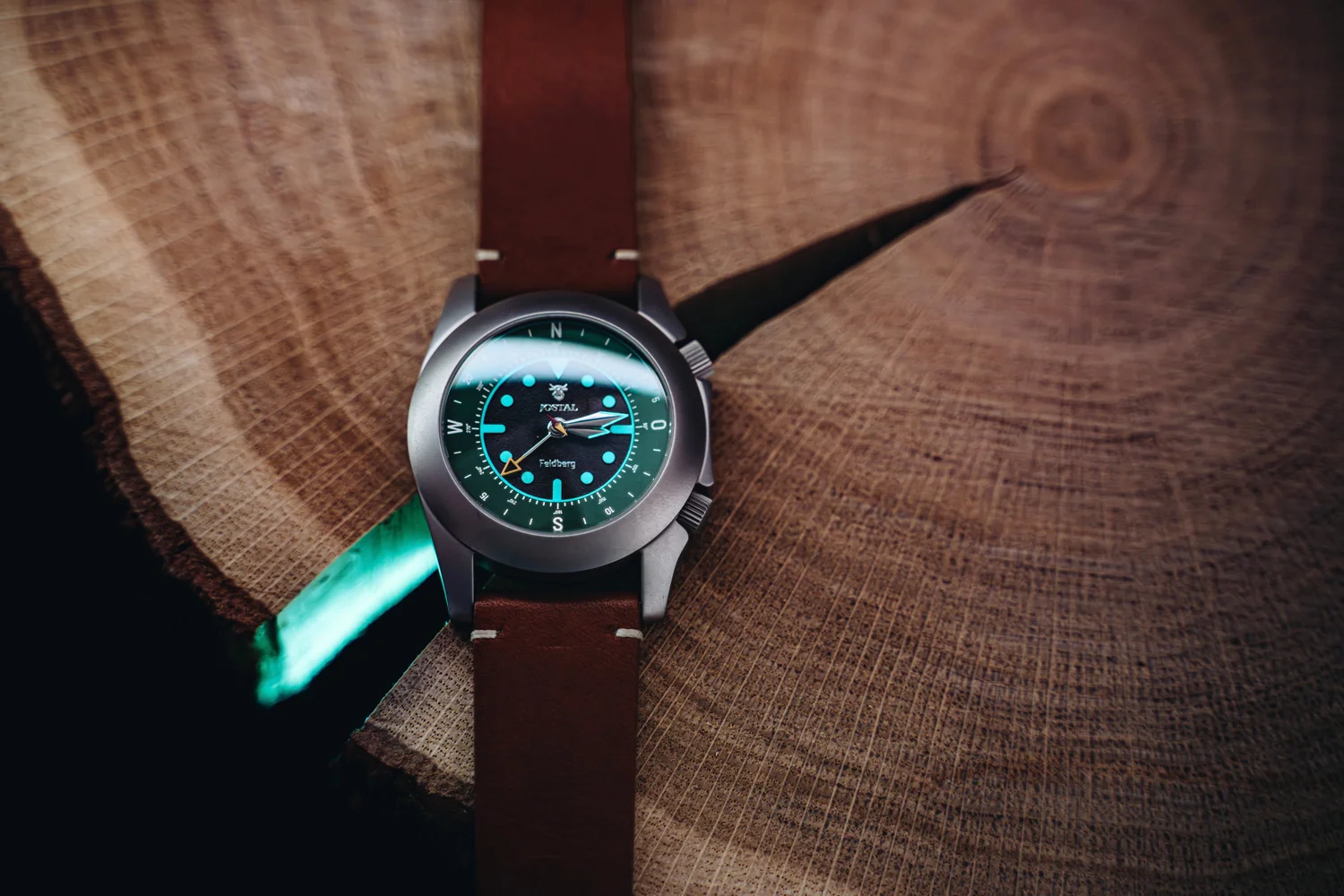 Grey Jostal Feldberg watch with glowing aqua dial and brown leather strap on a rustic wooden slab green crack.