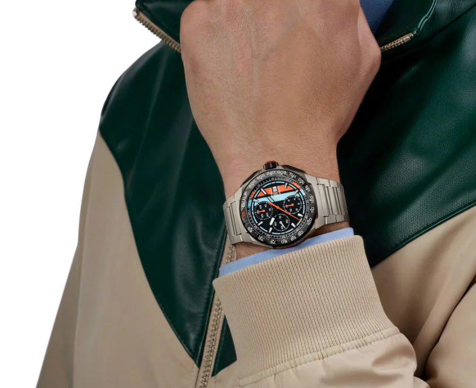 Person's wrist with steel chronograph watch, black dial, orange/blue Gulf stripes. Wears green & beige jacket, blue shirt cuff.