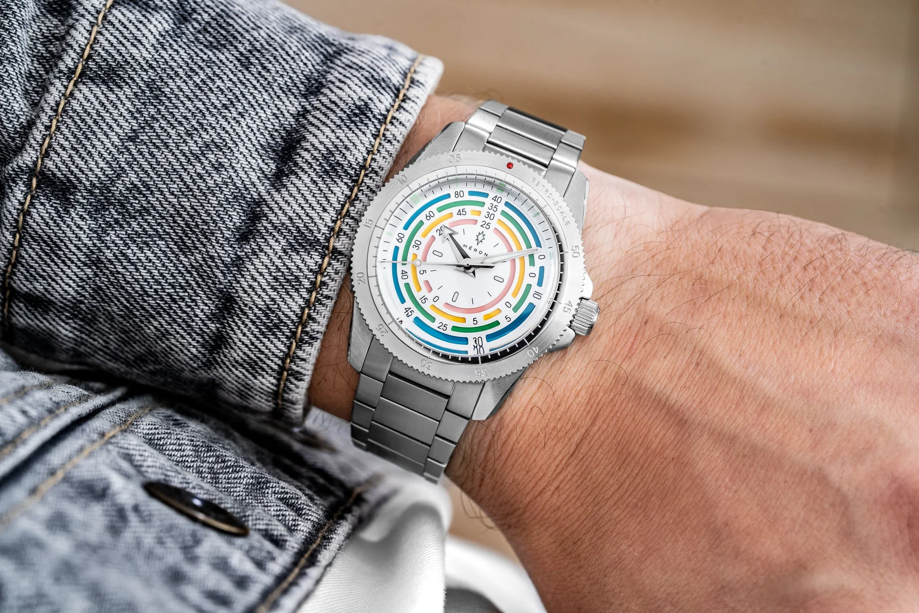 Wrist with a Heron watch: silver case, serrated bezel, metal bracelet, white dial with colorful concentric rings. Denim jacket.