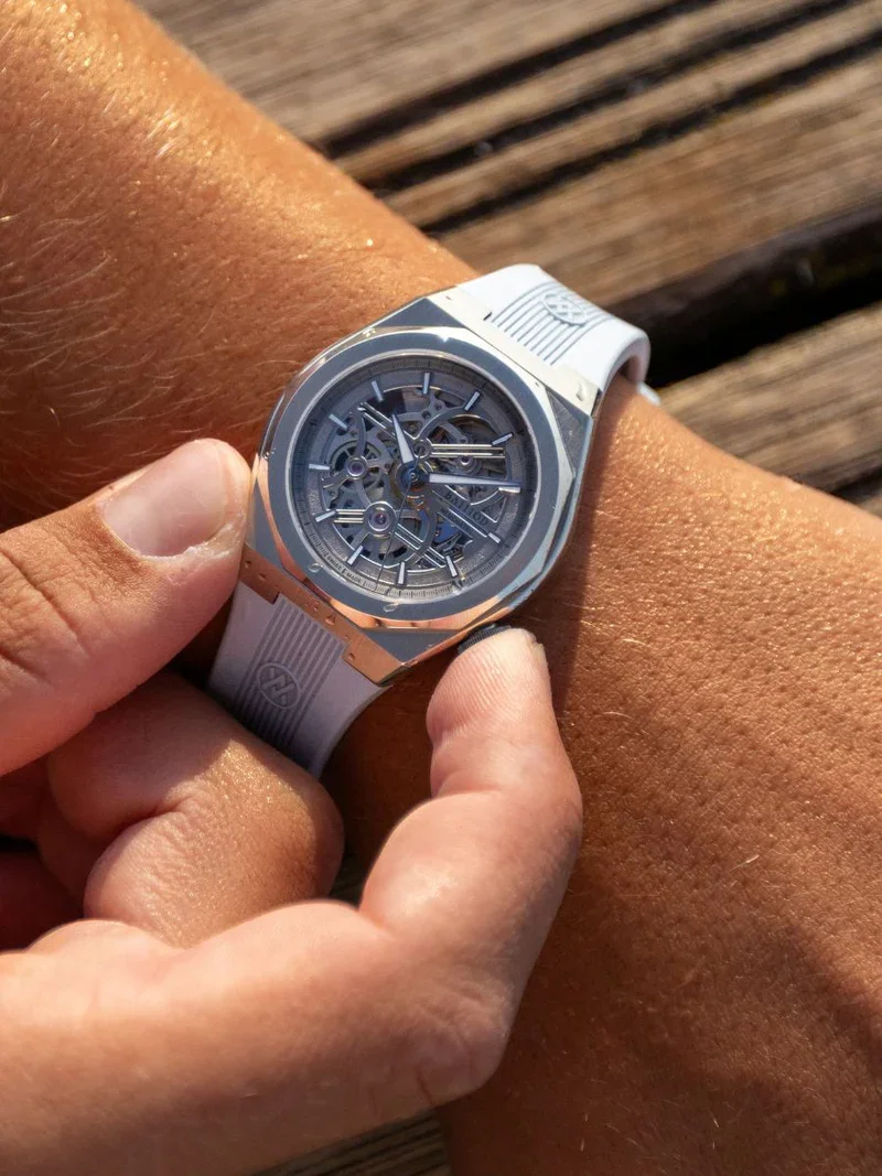 Tanned arm wears a luxury BYBLOS skeleton watch with silver case, rose gold accents & white rubber strap. Hand touches watch on wooden deck.