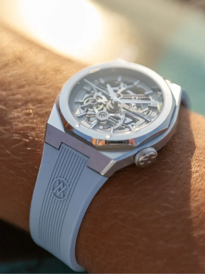 Close-up of a sunlit BA111OD Chapter 7 skeleton watch with an angular silver case and ribbed frost blue rubber strap on a wrist.
