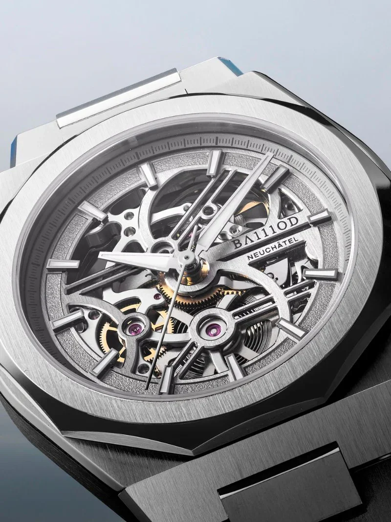 Close-up of a brushed silver BA111OD NEUCHÂTEL skeleton watch, revealing intricate gears and rubies through its open dial.