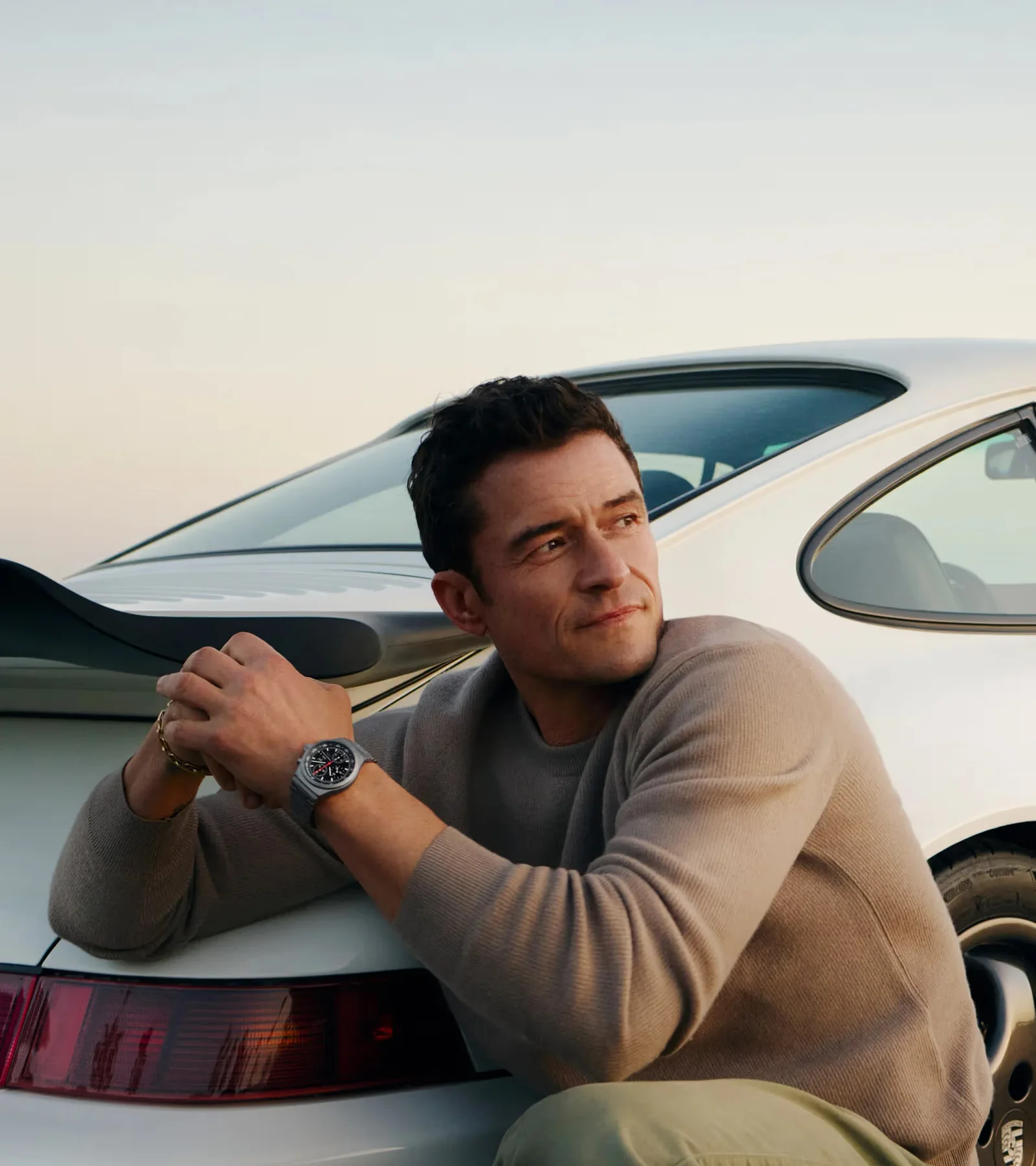 Orlando Bloom leans on a white Porsche 911 while wearing the Porsche Design Chronograph 1 All Titanium Numbered Edition.