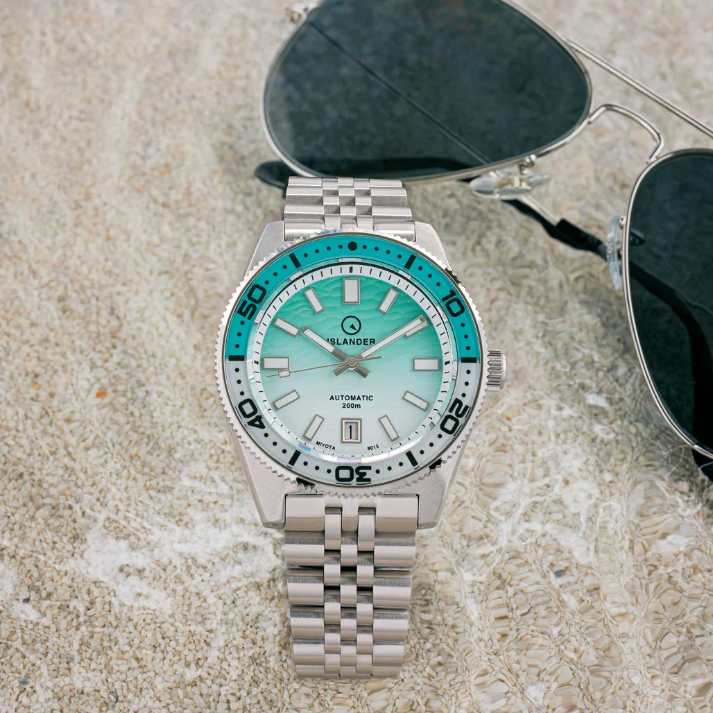 Islander automatic diver watch with teal bezel, gradient wave dial, steel bracelet, and aviator sunglasses on wet sand.
