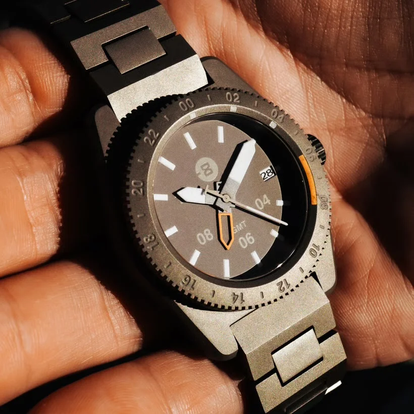 Close-up: Brushed titanium GMT watch on a hand. Dark grey dial, white markers, orange GMT hand, date 28.