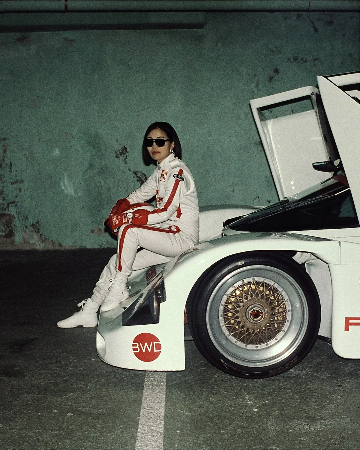 Woman in white TAG Heuer racing suit & shades sits on a white race car with gullwing door & gold wheel in a garage.